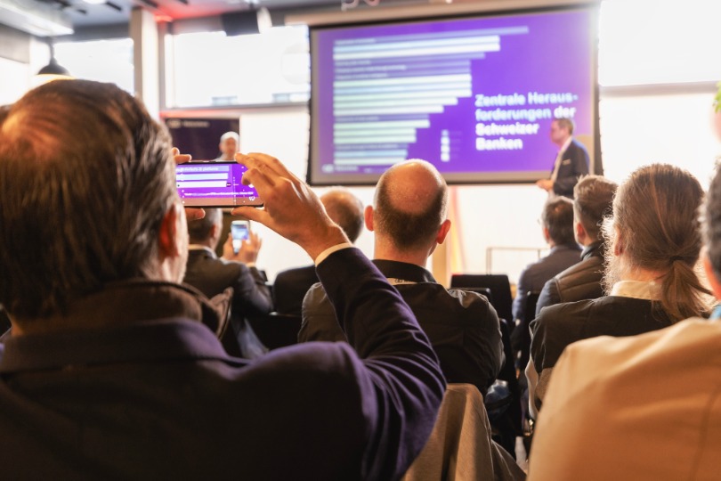 Audience members at a conference photographing a presentation slide showing key challenges for Swiss banks, with a speaker on stage. 