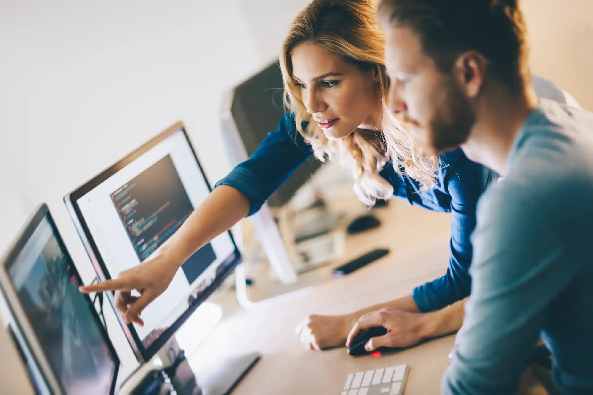 Woman pointing at a computer screen while discussing code with a colleague, illustrating collaboration on digital solutions.