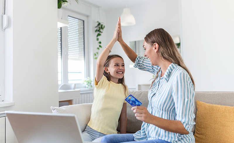 low-angle-excited-small-daughter-giving-high-five-mother-screaming-while-celebrating-successful-online-shopping-using-laptop-table-light-living-roomb