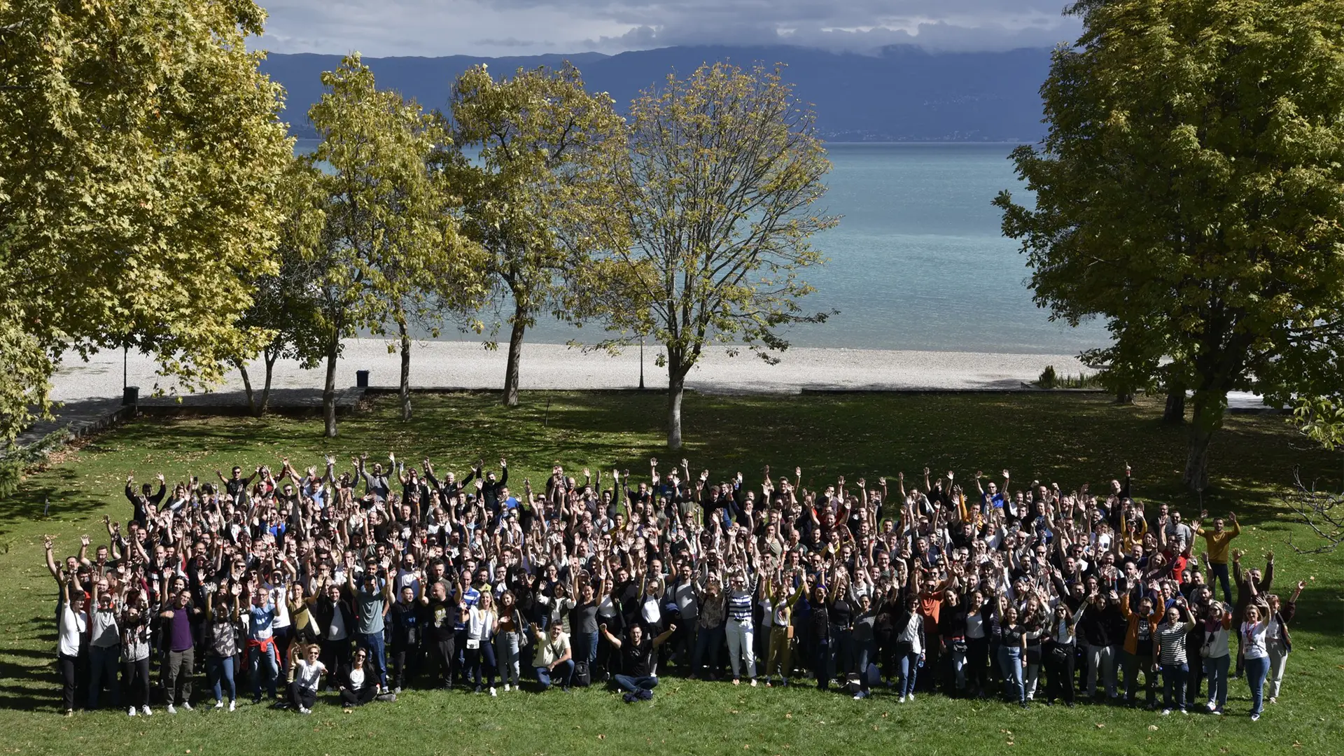 Large group of G+D Netcetera employees gathered outdoors by a lake, raising their hands in celebration, with trees and mountains in the background. 